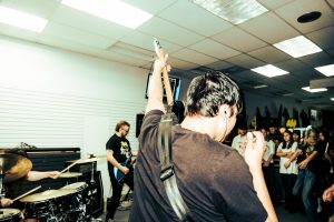 photo from behind of two men with guitars and a drummer performing for a small crowd