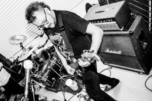 black and white photo of a man with a guitar in front of a stack of amps and standing at a microphone