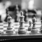 A black and white photo of a wooden chess board. The white bishops, queen, king, one knight, and several pawns are in focus.