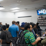 A group of young men standing around, waiting for a video game tournament to begin in the Mystery Shop, Oxnard, CA. Televisions mounted near the ceiling display the text "Let's Rock."