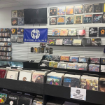 A collection of records hangs in display frames on the upper section of a wall. Below that, vinyl records are on display for sale on the wall and in record flip bins. There is a sign taped to the flip bin that says "GNG - Used Jackets Black or White, 10 for $5.00."