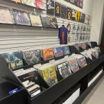 Various vinyl records on display in a shop. Some are on the wall and some are in record flip bins. A t-shirt also hangs on the wall on a clothes hanger.