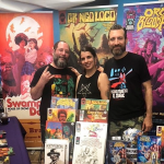 Two men and a woman pose, smiling, in front of three comic book banners. A title on one of the banners is partially obscured; the other two banners read "Gringo Loco" and "Orgy of the Blood Freaks." A selection of comic books is available for purchase on the table in front of them.