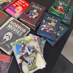 A collection of books and comic books laying on a table. The covers read "Jojo's Bizarre Adventure," "Destroy All Humans," "The Definitive Guide to Horror Movies," and "How to Kill a Guy in Ten Dates."