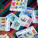 A picture of the cards for Loteria de Oxnard, scattered on top of a table with a colorful tablecloth and a bowl of popcorn in the bottom left corner.